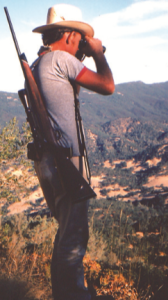 The author stands with a gun over his shoulder and binoculars to his eyes.