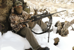 Young man in full winter hunting gear with rifle, sitting in the snow.